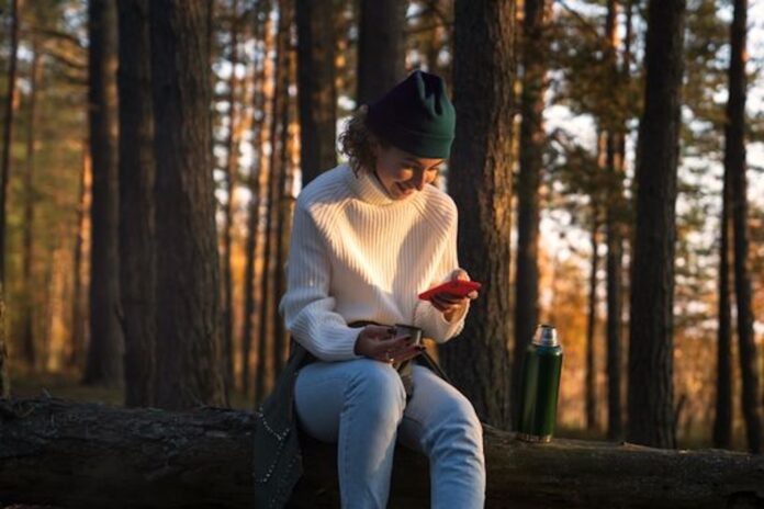 Kvinde i en hvid sweater sidder på en træstamme i skoven. Hun kigger på sin telefon imens hun smiler